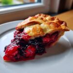 A juicy slice of triple berry pie resting on a white plate, showing the rich berry filling and flaky crust.