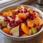 Close-up of a white bowl filled with a colorful winter fruit salad featuring orange segments, diced pears, and bright red pomegranate seeds.