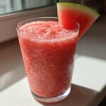 A tall glass filled with a bright red, icy watermelon smoothie, garnished with a fresh watermelon wedge.