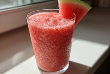 A tall glass filled with a bright red, icy watermelon smoothie, garnished with a fresh watermelon wedge.