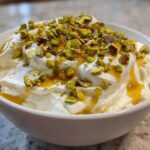 A white bowl filled with creamy whipped goat cheese, drizzled with honey and topped with chopped pistachios.