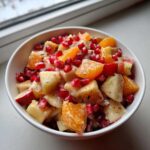 Close-up of a bowl filled with a colorful winter fruit salad featuring diced apples, orange segments, and bright pomegranate seeds.