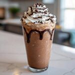 A tall glass filled with a thick, blended frozen hot chocolate topped with whipped cream, chocolate syrup drizzle, and shavings.