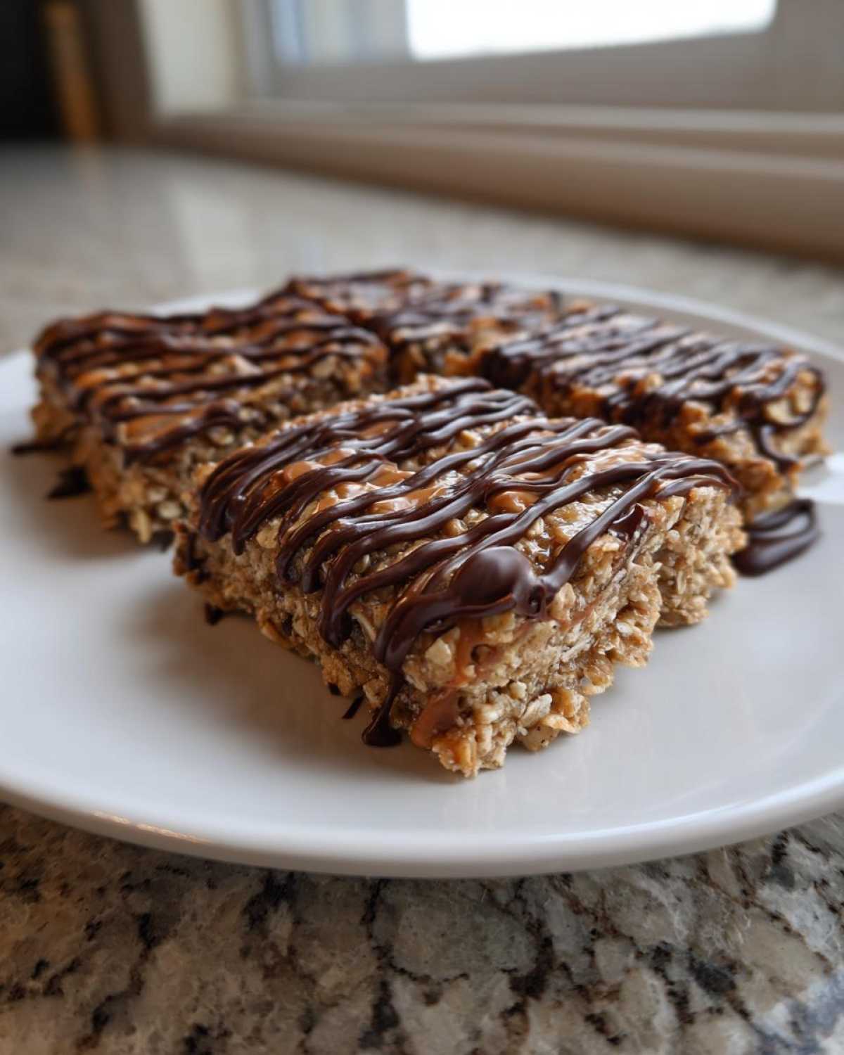 Close-up of four homemade protein bars drizzled with caramel and dark chocolate on a white plate.