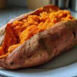 A close-up of a perfectly baked sweet potato, split open to show the fluffy, orange interior seasoned with pepper.