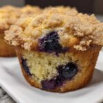 A close-up view of a moist blueberry muffin featuring visible blueberries and a thick, sugary crumb topping.