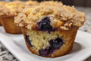 A close-up view of a moist blueberry muffin featuring visible blueberries and a thick, sugary crumb topping.