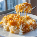 A spoonful of creamy, cheesy rice is lifted, showing long, stretchy strings of melted cheddar cheese from the cheesy rice dish.