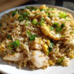 Close-up of amazing chicken fried rice piled high on a white plate, featuring pieces of chicken, scrambled egg, and green onions.