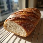 A loaf of golden-crusted French bread, sliced open to show the airy interior, resting on a wire cooling rack.