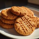 A stack of golden brown peanut butter cookies featuring classic crisscross fork marks, sitting on a white plate.