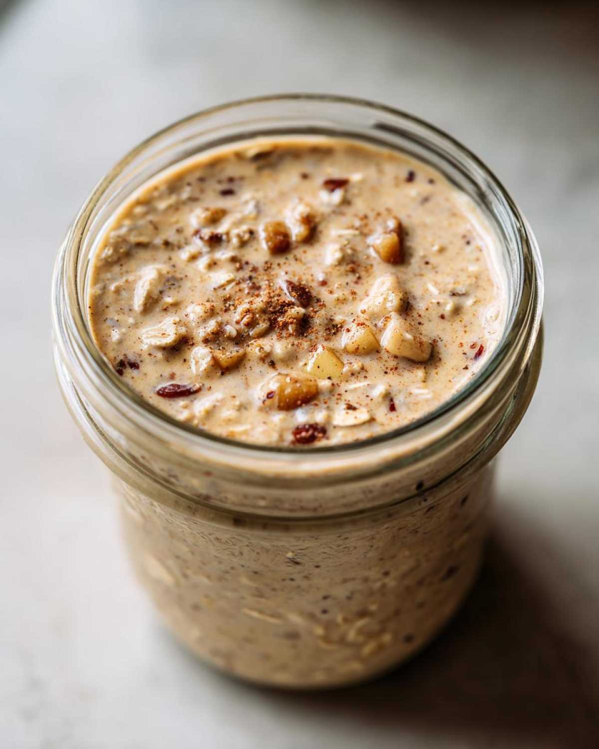 Overhead close-up of apple cinnamon overnight oats in a glass jar, topped with cinnamon and chopped nuts.