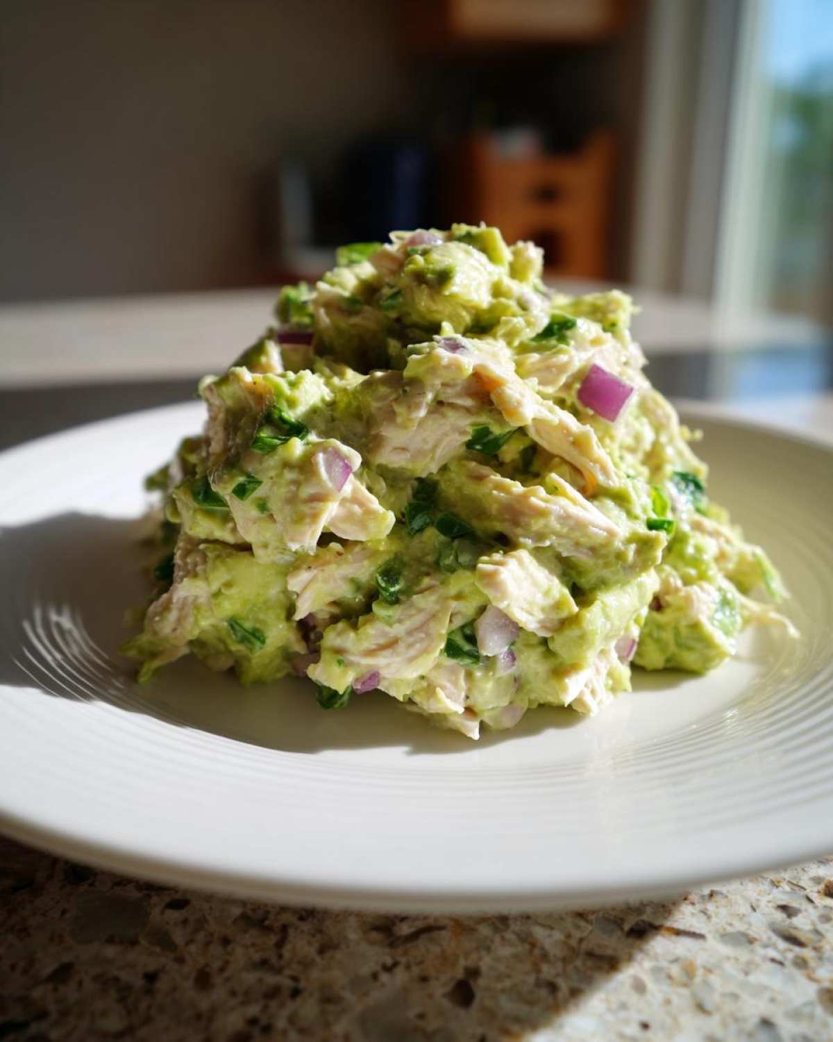 A mound of creamy avocado chicken salad mixed with red onion and green herbs served on a white plate.