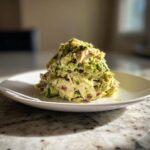 A mound of creamy avocado chicken salad featuring shredded chicken and visible chunks of red onion on a white plate.
