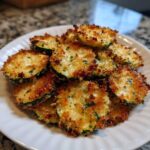 A pile of golden brown, crispy baked parmesan zucchini slices seasoned with herbs on a white plate.
