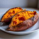 Two halves of a perfectly baked sweet potato showing bright orange, fluffy interior on a white plate.