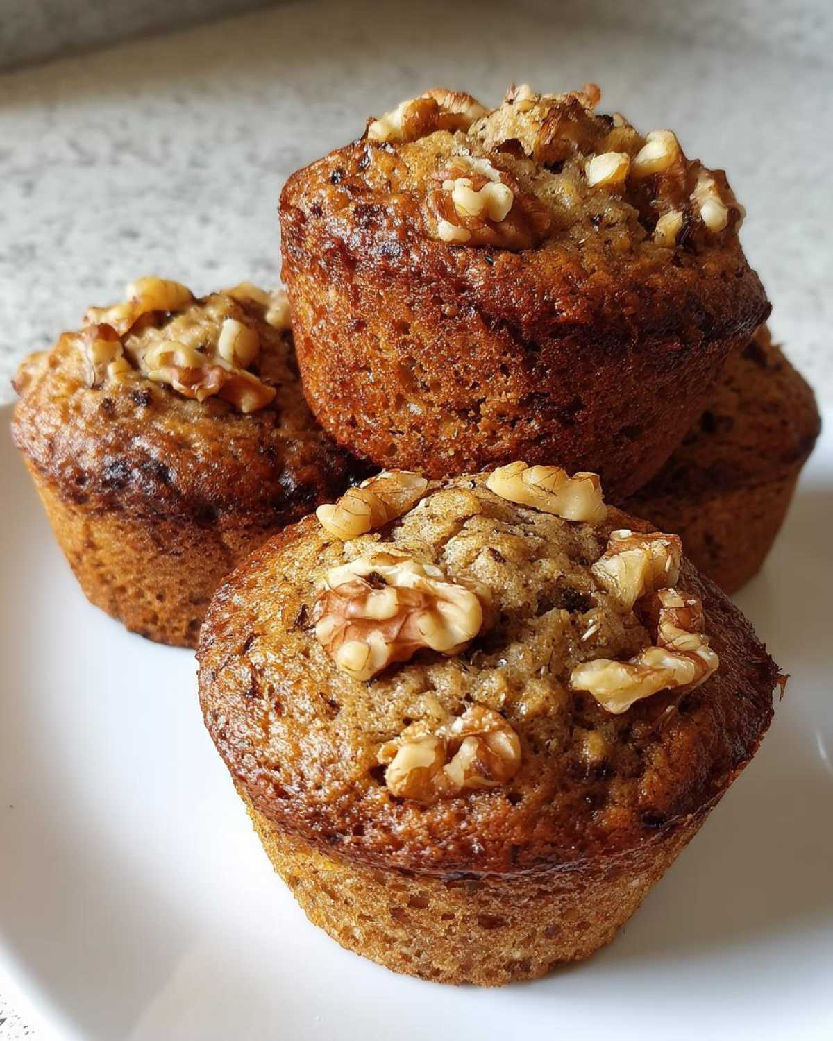 A stack of four golden brown banana nut muffins topped with crunchy walnuts on a white plate.