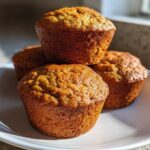 Four golden brown banana protein muffins stacked slightly on a white plate, highlighted by natural sunlight.
