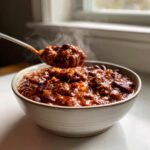 A spoonful of steaming, rich, meaty chili is lifted from a ceramic bowl, showcasing the best chili recipe ingredients.