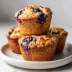 A close-up stack of three golden-brown blueberry protein muffins sitting on a light ceramic plate.