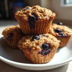 A stack of four golden-brown blueberry protein muffins with a crumb topping resting on a white plate.