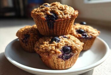 A stack of four golden-brown blueberry protein muffins with a crumb topping resting on a white plate.