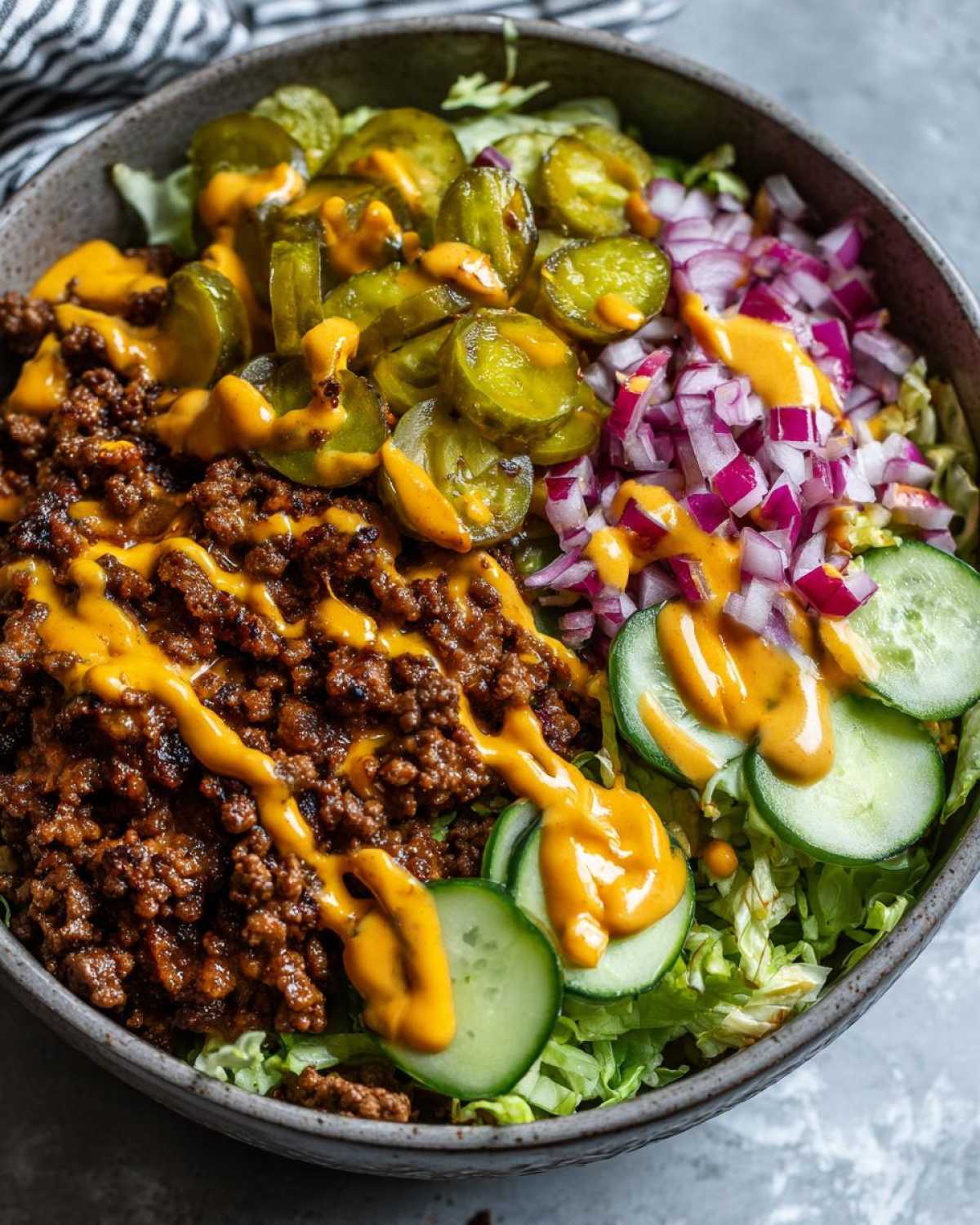 Close-up of a delicious cheeseburger bowl with seasoned ground beef, cheese sauce, pickles, and red onion over lettuce.