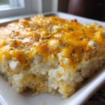 A square serving of baked cheesy chicken and rice casserole topped with melted cheddar and herbs.