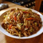 A close-up of a large white bowl filled with savory chicken chow mein recipe, featuring noodles, chicken pieces, cabbage, and carrots.