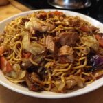 A close-up of a white bowl filled with savory chicken chow mein recipe, featuring noodles, chicken pieces, cabbage, and carrots.