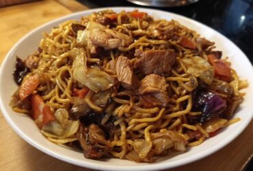 A close-up of a white bowl filled with savory chicken chow mein recipe, featuring noodles, chicken pieces, cabbage, and carrots.