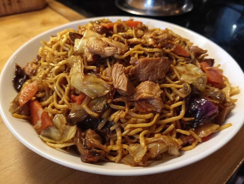 A close-up of a white bowl filled with savory chicken chow mein recipe, featuring noodles, chicken pieces, cabbage, and carrots.