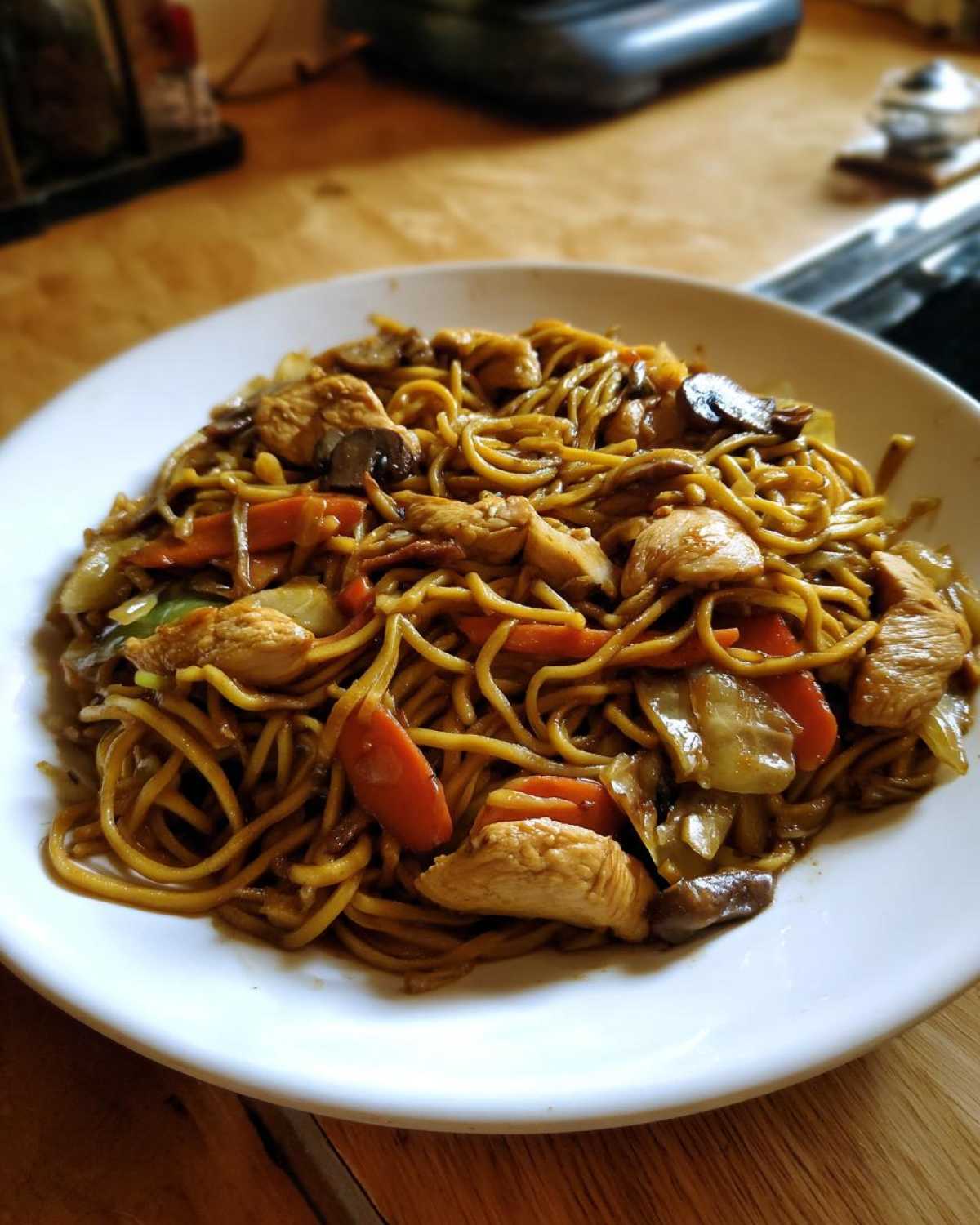 A close-up of a white plate filled with savory chicken chow mein recipe, featuring noodles, chicken pieces, carrots, and mushrooms.