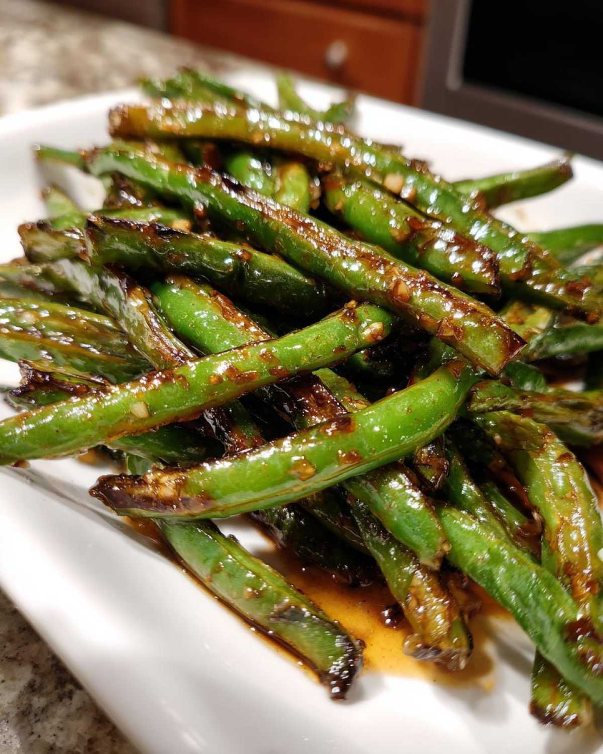 Close-up of bright green beans coated in a glossy, dark Chinese green beans with garlic sauce, served on a white plate.