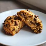 Two halves of a moist, golden-brown breakfast cookie packed with chocolate chips, resting on a white plate.