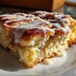 A close-up, sunlit shot of a gooey slice of Cinnamon roll casserole topped with thick white icing.