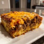 A close-up view of a hearty slice of cowboy cornbread casserole featuring a layer of seasoned ground beef topped with melted cheddar cheese.