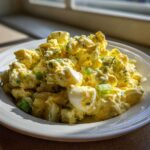 A white bowl filled with creamy, yellow egg salad recipe, featuring chunks of egg and green celery, highlighted by natural light.