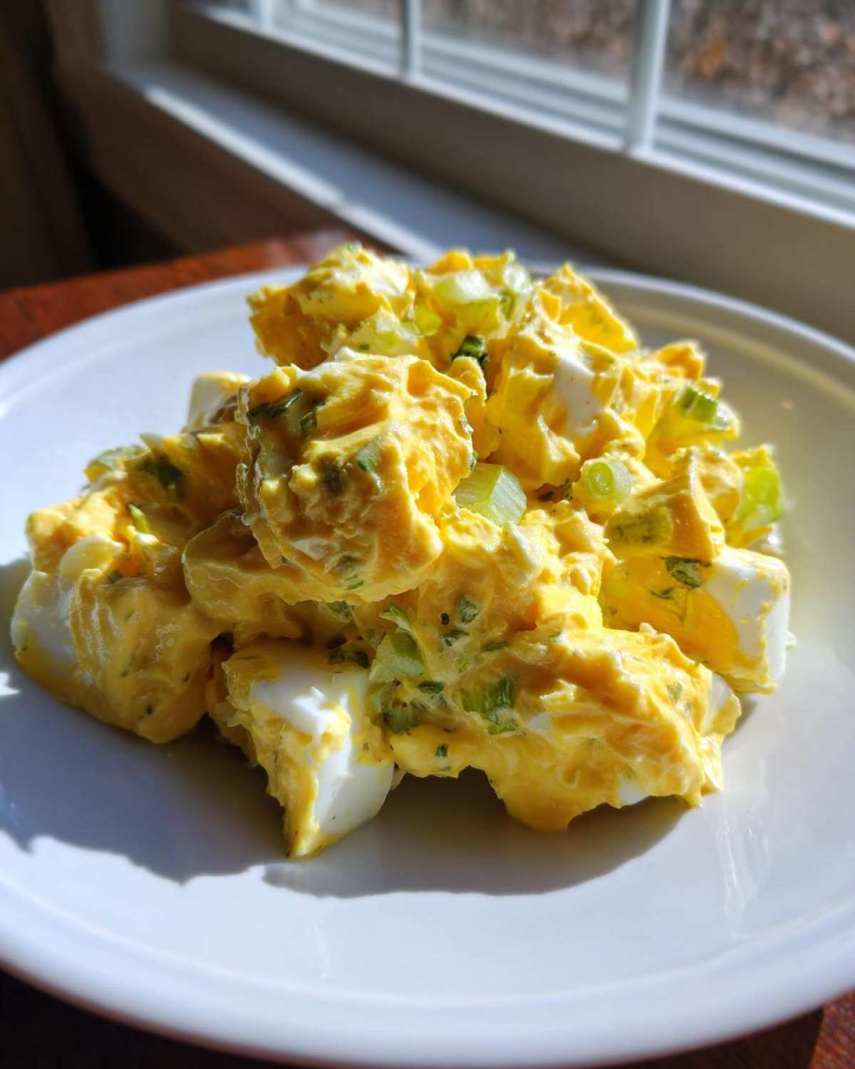 A close-up of creamy, chunky egg salad recipe mixed with celery and herbs, served on a white plate near a window.