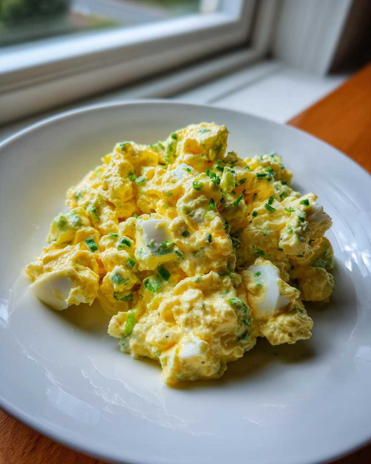 A close-up of creamy egg salad recipe, featuring chopped eggs and green chives, served on a white plate near a window.