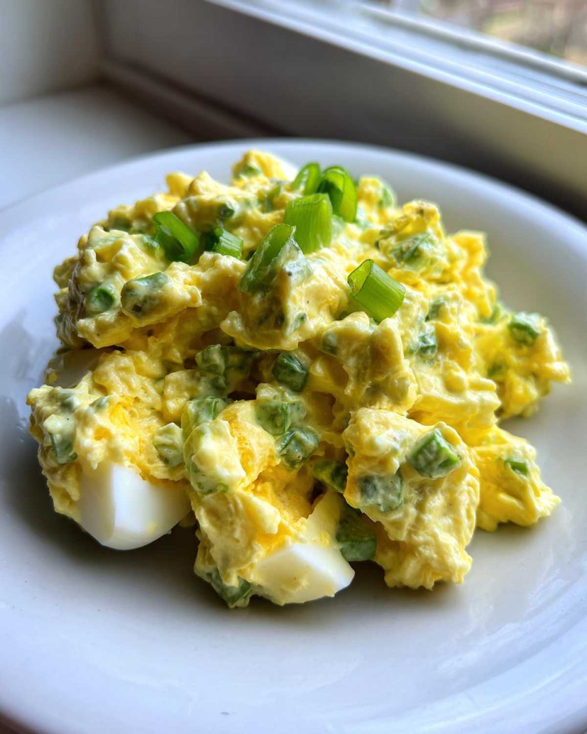 A close-up of creamy egg salad recipe mixed with green onions, served on a white plate near a window.