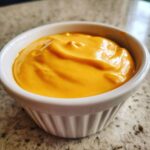 Close-up of smooth, bright orange homemade nacho cheese sauce served in a small white ramekin.