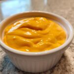 Close-up of vibrant orange, creamy nacho cheese sauce filling a small white ramekin on a speckled countertop.
