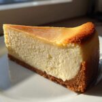 A perfect slice of creamy Protein Cheesecake with a graham cracker crust, beautifully lit by sunlight.