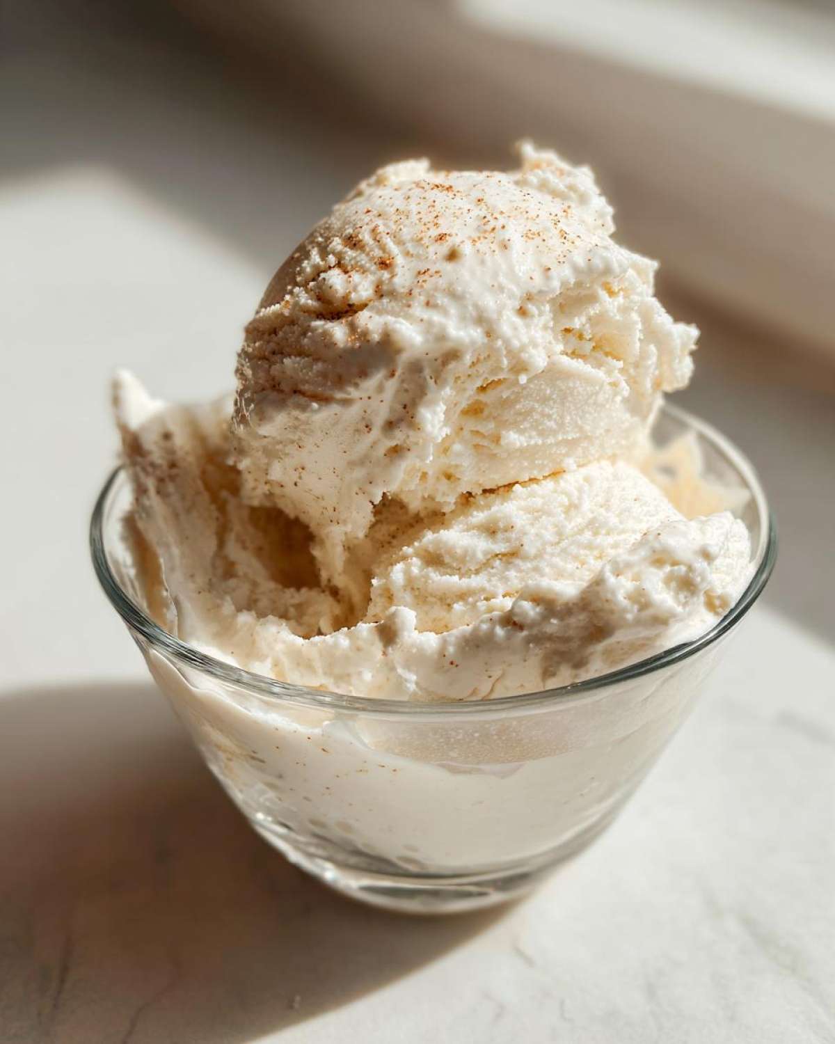 Two scoops of creamy, vanilla-colored protein ice cream sprinkled with cinnamon in a clear glass bowl.