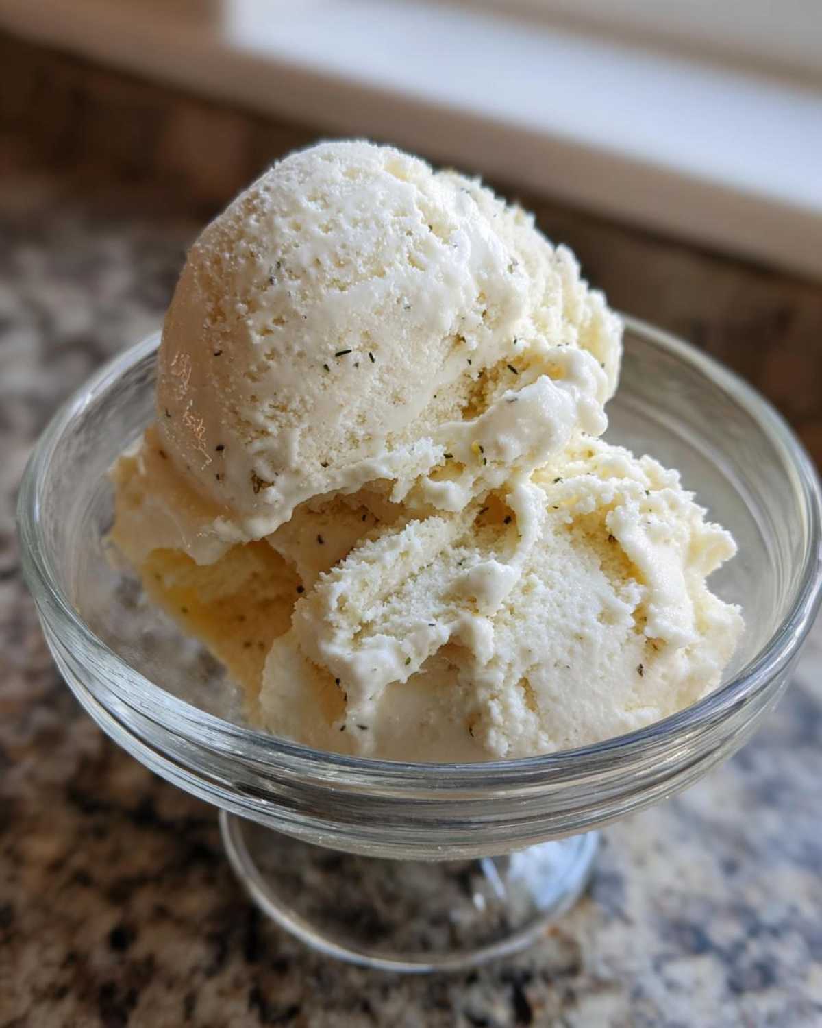 Close-up of two scoops of creamy, vanilla-colored protein ice cream speckled with dark flecks in a clear glass dish.
