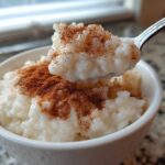 A spoonful of creamy rice pudding topped with ground cinnamon being lifted from a white bowl.