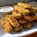 A tall stack of golden brown, crispy Fried Pickles served on a white plate next to a small ramekin of dipping sauce.