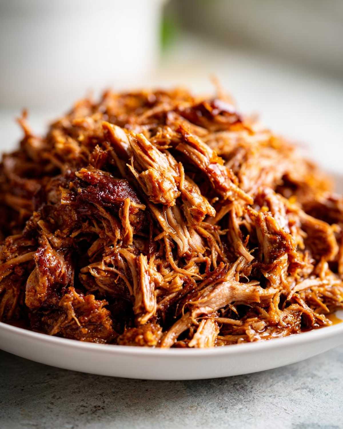 A close-up shot of tender, saucy crock pot pulled pork piled high on a white plate.