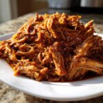 A close-up of tender, saucy crock pot pulled pork piled high on a white plate, glistening in the sunlight.